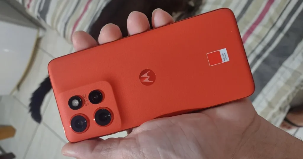 Uma pessoa segurando na palma da mão um smartphone Motorola Edge 50 Neo na cor Pantone Peach Fuzz (um tom de pêssego). A imagem foca na parte traseira do aparelho, mostrando o módulo de câmeras e o selo da Pantone.