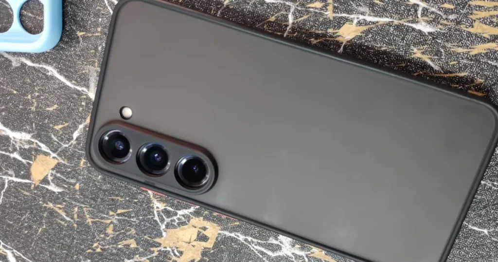 Close-up da traseira de um smartphone Samsung Galaxy dentro de uma capa preta. O módulo com as três câmeras verticais e o flash está em destaque sobre uma superfície com estampa de mármore.