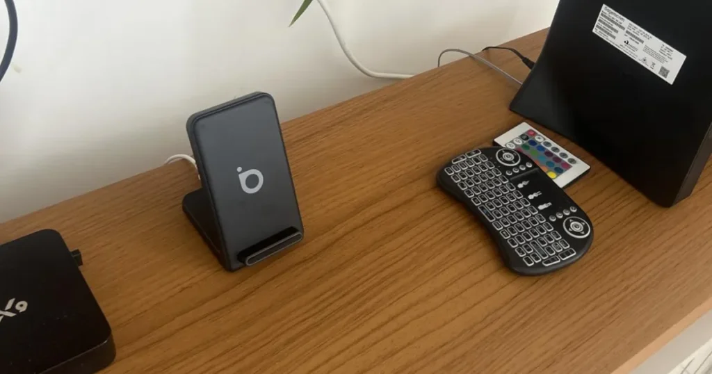 Um carregador sem fio do tipo suporte está sobre um móvel de madeira, ao lado de um mini teclado sem fio e outros aparelhos eletrônicos, como um roteador e um conversor de TV.