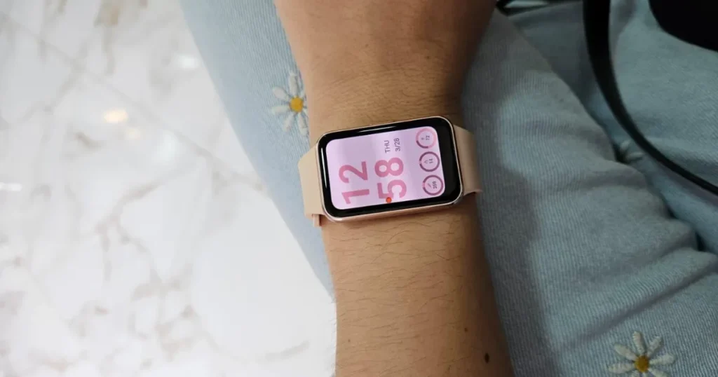 Uma pessoa usando no pulso a pulseira inteligente Samsung Galaxy Fit3 com corpo rosê e pulseira rosa-claro. A tela do dispositivo está ligada, mostrando um mostrador de relógio digital.