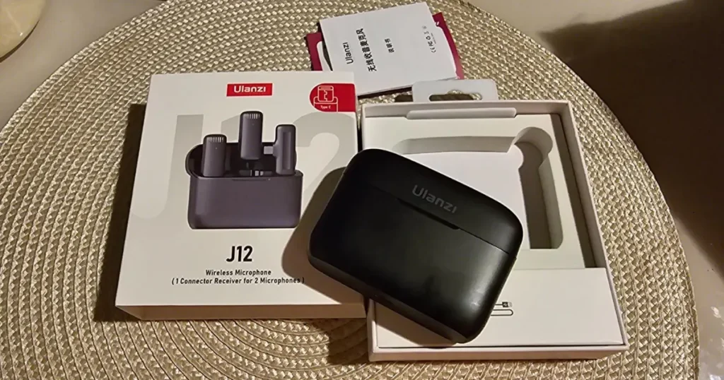 Unboxing do microfone sem fio Ulanzi J12. A caixa do produto está ao lado do estojo de carregamento preto sobre uma mesa com sousplat de tecido.