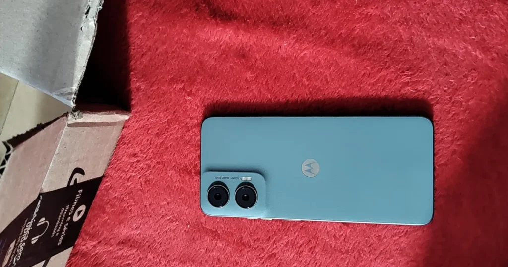 Fotografia de um celular falsificado, imitando um conceito do "Moto G35". O aparelho, na cor verde-água, está deitado sobre uma superfície vermelha ao lado de uma caixa de papelão.