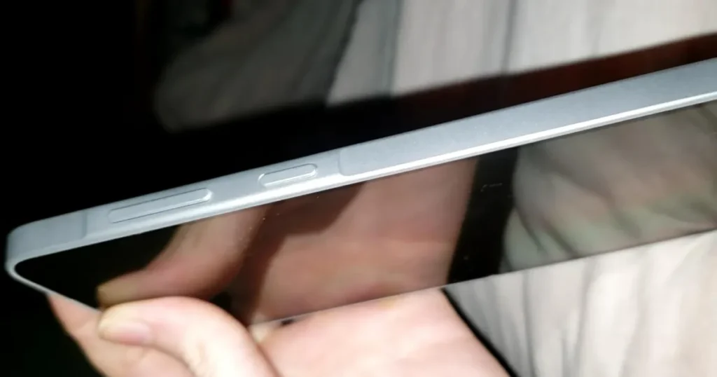 Fotografia em close-up da lateral de um smartphone com moldura prateada, mostrando em detalhe os botões de volume e de energia. O aparelho está sendo segurado por uma mão.