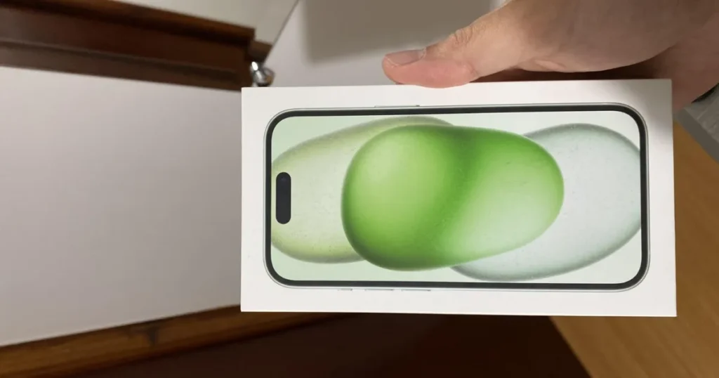 Pessoa segurando uma caixa de iPhone 15 com a imagem do aparelho na cor verde na parte frontal da embalagem. A caixa está inclinada na horizontal, em ambiente interno.