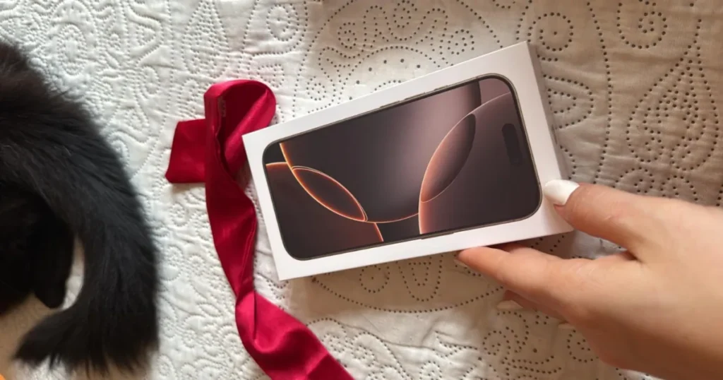 Caixa de um iPhone em cima de uma colcha clara, sendo segurada por uma mão com unhas pintadas de branco. Há uma fita vermelha e parte de um gato preto na imagem.