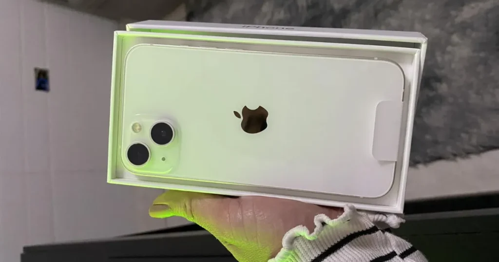 Uma pessoa segurando a caixa aberta de um iPhone 13 branco (cor Estelar), exibindo a parte de trás do aparelho novo com o plástico protetor e o recorte para o logo da Apple e o módulo de câmera dupla.